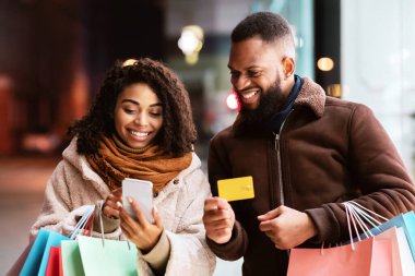 İnternetten sipariş veriyorum. Gülümseyen siyah bir çift cep telefonu kullanıyor, bankamatik kartı gösteren bir adam, elinde renkli alışveriş çantaları olan bir aile, akşamları dışarıda dolaşıyor, internetten alışveriş yapıyor.