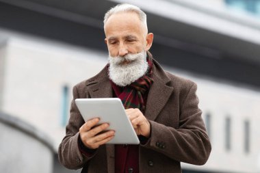 Zengin kıdemli gri saçlı şık giyimli adam dijital tablet kullanıyor, şehir içinde yürürken konumu kontrol ediyor, harita kullanıyor ya da internet üzerinden ziyaret edecek yer arıyor, uzay kopyaları yapıyor.