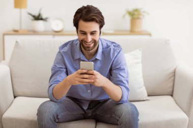 Kulaklıklı mutlu adam evdeki koltukta oturan akıllı telefondan video görüşmesi yapıyor. Oturma odasında telefonla mesajlaşan adam. Teknoloji ve Gadget, Harika Mobil Uygulama Konsepti