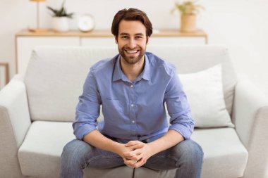Kanepede oturan ve evdeki kameraya gülümseyen yakışıklı adam. Başarılı Millennial Serbest Çalışan Adam Salonda Poz Veriyor. Kendinden emin Erkek Portresi Konsepti