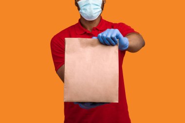 Kilitleme sırasında teslimat servisi. Tanımlanamayan Afro-Amerikan erkek kurye, tek kullanımlık maske takıyor ve elinde içi boş kağıt paketler tutan koruyucu eldivenler tutuyor.