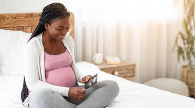Bebek ultrason görüntüsüyle yatakta oturan mutlu Afrikalı hamile kadın cenin sonografi fotoğrafına bakıyor, siyah kadın sağlıklı hamileliğinin ve gelecekteki anneliğinin tadını çıkarıyor,