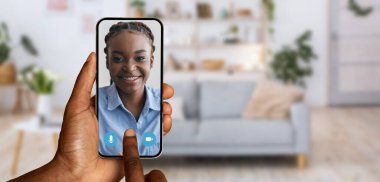 Bir kişi akıllı telefon kullanarak bir video görüşmesine katılıyor, ekranda neşeli bir gülümseme gösteren bir kadın. Arka planda rahat bir oturma odası var. Ev ortamını güzelleştiriyor..