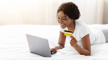 Evden online alışveriş, e-ticaret konsepti. Mutlu Afro-Amerikalı kadın evde yatıp internetten kıyafet ya da ürün alıyor. Kredi kartıyla ödeme yapıyor.