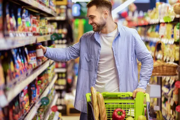 Her gün alışveriş konsepti. Gülümseyen genç adam markette koridorlar arasında tramvay sürüyor. Pozitif adam yiyecek alıyor, raftan ürün alıyor. Tüketim ve Yaşam Tarzı