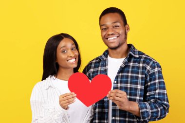 Sevgililer günü, yıldönümü ve modern ilişki. Gülümseyen bin yıllık Afro-Amerikan erkeği ve hanımefendisi, kırmızı kalbi kendi başına, sarı arka planda, stüdyo çekimlerinde, kopyalama alanında tutarlar.