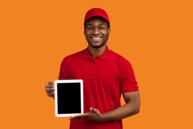 Teslimat Konsepti. Gülümseyen siyah kurye portresi elinde boş ekran ile dijital tableti tutuyor ve kamera önünde kırmızı başlık takıyor ve üniforma giyiyor turuncu arka planda tek başına duruyor.
