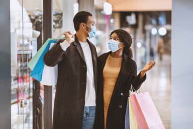 Kara Cuma ve sezonluk satışlar sırasında inanılmaz indirimler ve düşük fiyatlar. Şaşkın, Afro-Amerikalı, koruyucu maskeli kadın ve erkek alışveriş merkezinde bir sürü alışveriş torbası tutuyor.