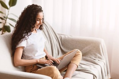 Koyu kıvırcık saçlı, evden çalışan, koltukta oturan, dizüstü bilgisayar kullanan, internet üzerinden mesaj yazan ya da müşterilerle iletişim kuran, blog yazan ya da kopyalayan genç bir kadın.