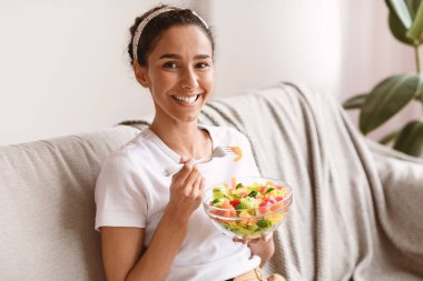 Gülümseyen güzel bayanın elinde taze salata ve çatalla evdeki kanepede oturup fotokopi çekmesi. Sağlıklı genç esmer kadın öğle yemeğinde sebze yiyor. Diyet kavramı