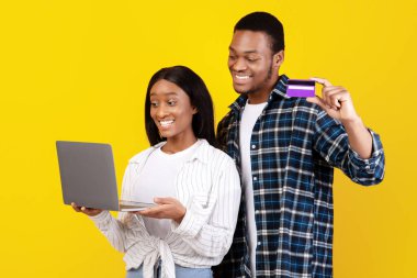 Alışveriş kolikleri, online alışveriş ve başarı duyguları. Gülümseyen bin yıllık Afro-Amerikan öğrenciler dizüstü bilgisayarla ödeme yapan ve sarı arka planda izole edilmiş kredi kartı gösteren karı-koca.