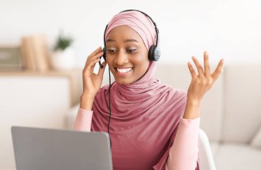 Online eğitim. Genç siyahi kadın öğretmen tesettürde öğrencilerle video dersi alıyor, evde dizüstü bilgisayar kamerası kullanıyor. Türbanlı mutlu Müslüman hoca evden giysi getiriyor.