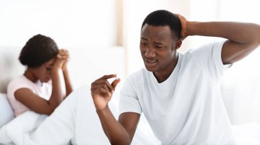 Ereksiyon bozukluğu, ilişkilerde ya da evlilik kavramında cinsel sorunlar. Kafası karışmış siyahi bir adam elindeki hapa bakıyor ve kafasına dokunuyor. Afro-Amerikan bir kadın yatakta arka planda oturuyor.