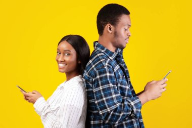 İlişkiler, sosyal ağlar ve internette sörf. Genç Afro-Amerikan erkek ve kadın telefonlarla sırt sırta dururlar, kadın gülümser ve kameraya bakar, sarı arka planda izole bir şekilde.