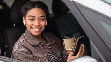 Cihazlar ve kahveyle mola vermiş modern bir sürücü. Mutlu Afrikalı genç Amerikalı bayan elinde bir fincan sıcak içecek paketiyle arabada oturuyor, akıllı telefonlu kablosuz kulaklıklar takıyor ve kameraya bakıyor, bedava uzay.