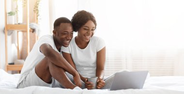 Yatakta oturan mutlu siyah çift, dizüstü bilgisayarda film izliyor, ev içi, fotokopi odası. Neşeli Afro-Amerikalı kadın ve erkek hafta sonunu birlikte geçiriyor, çay içiyor ve web sörfü yapıyor.