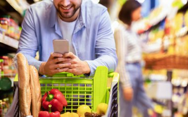 Süpermarkette yiyecek alışverişi yapan, hipermarkette Cart 'la ayakta duran, Mobil Kupon uygulaması kullanan tanınmayan adamın yakın çekimi. Gülümseyen Adam Dükkanda Akıllı Telefon Satın Alma Marketi Kullanıyor