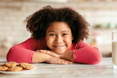 Çocukluğun kutlu olsun. Kurabiye ve sütle masada oturan sevimli siyah kız, lezzetli atıştırmalıklar yerken, kameraya bakıp gülümserken, neşeli Afrikalı kadın çocuk mutfakta poz veriyor.