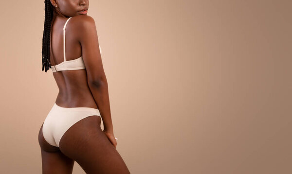 Обрезанный портрет молодой худой черной женщины в нижнем белье с растяжками на теле знаки Поза над бежевой студией фоне, позитивный африканская леди наслаждаясь своей природной красотой и несовершенство кожи
