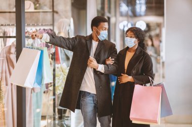 Kara Cuma 'da koronavirüs salgını ve sosyal mesafeler sırasında alışveriş merkezinde maske takan çiftlerin portresi. Milenyum Afrikalı Amerikalı adam butik vitrinini işaret ediyor.