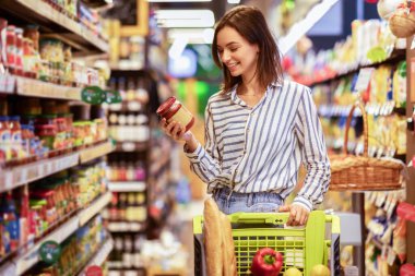 Süpermarkette alışveriş arabasıyla gülümseyen bir kadının portresi koridorda yürürken ve markette raflarda yürürken, cam sos kavanozu tutarken, alışveriş merkezinde sağlıklı ürünler seçerken.