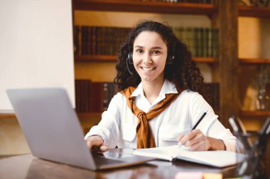 Karantina sırasında e-öğrenim ve uzak eğitim. İnternette çalışan, dizüstü bilgisayar kullanan, not alan, kameraya bakıp poz veren genç bir kız öğrenci.