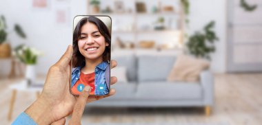 Bir kişi akıllı telefondan dostça bir video görüşmesi yapıyor, gülümseyen Arap kadınla sohbetin tadını çıkarıyor. Konforlu ev ortamında arka planda bir kanepe ve dekoratif öğeler yer almaktadır..