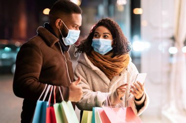 Perakende, Teknoloji ve Salgın Konsepti. Tıbbi maske takan, akıllı telefon kullanan, ekranı işaret eden ve elinde alışveriş torbaları olan kocasına uygulama gösteren Afrikalı Amerikalı bir çiftin portresi.