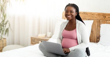 Hamile Kadınlar için İnternet 'te iş. Siyah Müstakbel Anne evde dizüstü bilgisayar kullanıyor, bilgisayarla yatakta oturuyor, bebek beklerken uzaktan çalışıyor, kameraya gülümseyen güzel Afrikalı Kadın