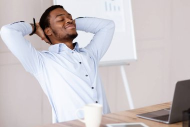 Modern ofisteki dizüstü bilgisayarının önünde oturan neşeli Afro-Amerikan genç menajeri başarılı bir iş anlaşmasından ya da internet konferansından sonra dinleniyor.