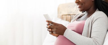 Neşeli siyahi hamile kadın evde akıllı telefonuyla vakit geçiriyor, internete bakıyor ya da arkadaşlarıyla mesajlaşıyor, Afrikalı beklenen kadın rahat yatağında dinleniyor, fotokopi çekilen kenar görüntüsü