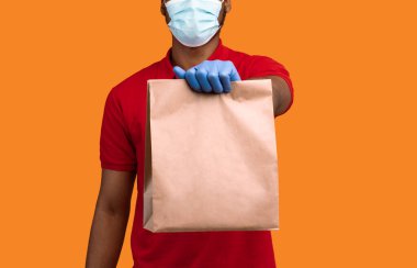 Salgın sırasında doğum. Cerrahi maske takan, koruyucu eldiven takan, kırmızı üniformalı ve tasarım için boş alanı olan el işi kağıt paketi veren Afrika kuryelerini kapatın.