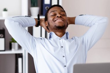 Modern ofisteki dizüstü bilgisayarının önünde oturan neşeli Afrikalı Amerikalı genç adam başarılı bir iş anlaşmasından ya da internet konferansından sonra dinleniyor.