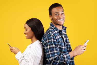 Online randevu, aşk, cihazlar ve sosyal ağ. Milenyum Afrikalı Amerikalı gülümseyen adam akıllı telefonu tut ve kameraya bak, telefonda yazan kadın, sarı arka planda izole edilmiş, stüdyo çekimi