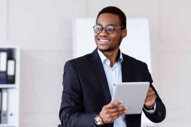 Modern aletler ve iş. Mutlu Afro-Amerikalı takım elbiseli genç iş adamı ofiste dijital tablet kullanıyor, internette çalışıyor, sunuma hazırlanıyor, fotokopi alanına bakıyor ve gülümsüyor.