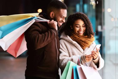 İnternetten kıyafet sipariş ediyorum. Afro-Amerikan çiftin portresi akıllı telefon kullanıyor, ellerinde aletler ve kağıt poşetler tutuyor, akşamları dışarıda dikiliyorlar. Perakende, Satış, Teknoloji