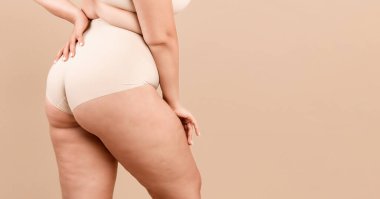 Artı Boyutlu Kadın. Tanınmayan kıvrımlı kadın iç çamaşırlı, büyük beden kadın iç çamaşırlı, bej renkli stüdyo arkasında duruyor, doğal güzelliğinin, panoramasının, bedava uzay boşluğunun tadını çıkarıyor.