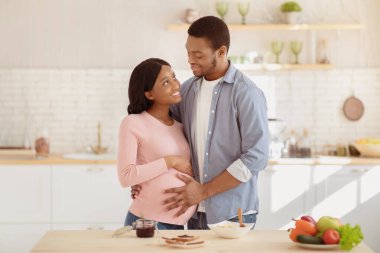Mutlu siyah adam, güzel hamile kız arkadaşına sarılıyor ve mutfakta karnına dokunuyor. Sevgili milenyum çifti evde sağlıklı yemek pişirirken kucaklaşıyorlar. Ebeveynlik kavramı