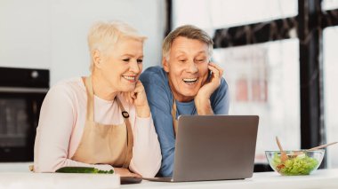 Son sınıfların eşleri modern mutfaktaki dizüstü bilgisayarın başında otururken çekilmiş videoları. Mutlu Emekli Yaşam Stili. Yaşlı bir çift internette birlikte yemek pişiriyor ve internette geziniyor..
