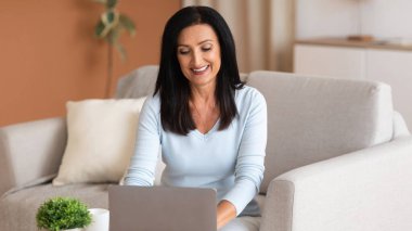 Teknoloji. Dahili ofiste uzaktan bilgisayarla çalışan çekici, olgun bir iş kadını uzaktan kumandalı işten zevk alan, masadaki bilgisayarla koltukta oturan, klavyeye yazı yazan bir kadın.