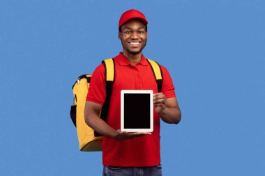 Modern Teknoloji ve Teslimat. Gülümseyen siyah erkek kurye elinde dijital tabletle kamera önünde kırmızı üniforma ve sarı termo çanta ile mavi stüdyoda duruyor.