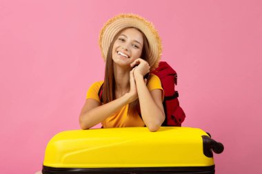 Büyük sarı bavula yaslanmış, kameraya gülümseyen, pembe stüdyo arka planı olan, reklam için fotokopi çeken, yaz şapkalı mutlu bir genç kadın. Neşeli bayan gezgin tatile çıkıyor.