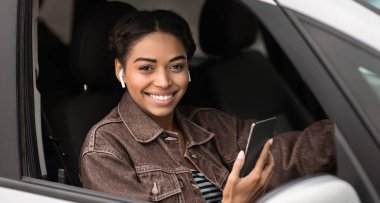 Şoförlük, modern taksi şoförü ve şehirde meslek. Kablosuz kulaklıklı gülümseyen Afro-Amerikan kadın. Elinde telefon, gümüş renkli arabada oturuyor, açık pencereden bakıyor, boşluğu kopyalıyor.