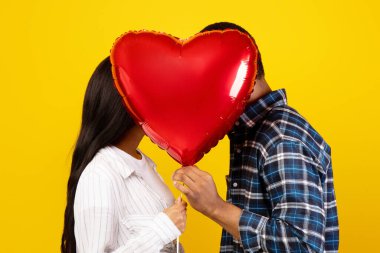 Sevgililer Günü 'nde romantik bir öpüşme ve randevu. Milenyum utangaç Afrikalı erkek ve kadın Coronavirus salgınında yüzü kalp balonuyla kaplanmış, sarı arka planda izole edilmiş, stüdyo çekimi, profil