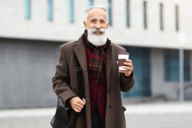 Caddede yürüyen, kağıt bardakta kahve içen, kameraya gülümseyen, fotokopi çeken mutlu yaşlı bir adam. Neşeli büyükbaba hafta sonu etrafı geziyor, sıcak çay içiyor, ısınıyor.