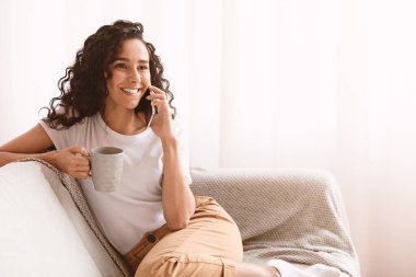 Ev kıyafetleri içinde siyah kıvırcık saçlı neşeli bir bayan akıllı telefondan konuşuyor, çay ya da kahve içiyor oturma odasında kanepede oturuyor, hafta sonları dinleniyor ve fotokopi odasına bakıyor.