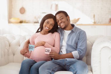 Kanepede oturan, ellerinde çocuk ve kız kartları tutan, gülümseyen ve evde kameraya bakan sevecen siyah çift. Genç Afrikalı Amerikalı ebeveynler doğmamış bebeklerinin cinsiyetini tahmin ediyorlar.