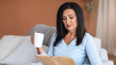 Evde vakit geçirmek. Yetişkin kadın portresi kitap okuyor ve elinde bir fincan kahve tutuyordu. Oturma odasındaki kanepede oturmuş hafta sonunun keyfini çıkarıyordu. Kadın dinleniyor ve dinleniyor.