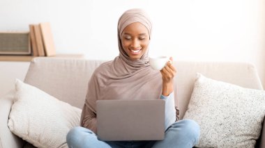 Keyifli siyah kadın tesettürde ailesiyle ya da arkadaşıyla bilgisayarda sohbet ediyor, rahat bir kanepede kahve içiyor. Müslüman kadın internet üzerinden iletişim kuruyor, web dersi alıyor ya da toplantı yapıyor