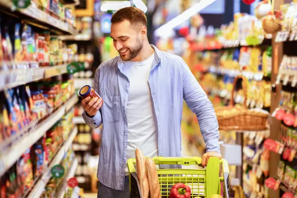 Tüketim Konsepti. Gülümseyen Sakallı Adam 'ın portresi markette alışveriş arabasıyla marketten yiyecek alıyor koridorda yürüyor ve raflarda duruyor, elinde cam kavanoz sos tutuyor, etiketi kontrol ediyor.